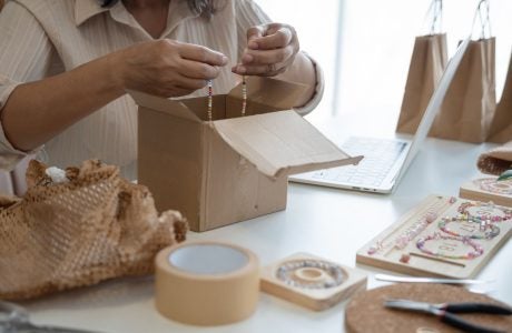 a woman who owns a jewelry small business is packaging a bracelet into a cardboard box on top of a table with lots of jewelry supplies and bags, she is fulfilling an online order.