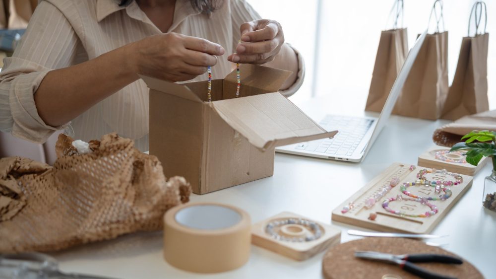 a woman who owns a jewelry small business is packaging a bracelet into a cardboard box on top of a table with lots of jewelry supplies and bags, she is fulfilling an online order.