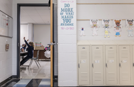 Student sitting at a desk in a classroom raises her hand, and a sign posted in the hallway says "Do more of what makes you awesome" next to lockers.