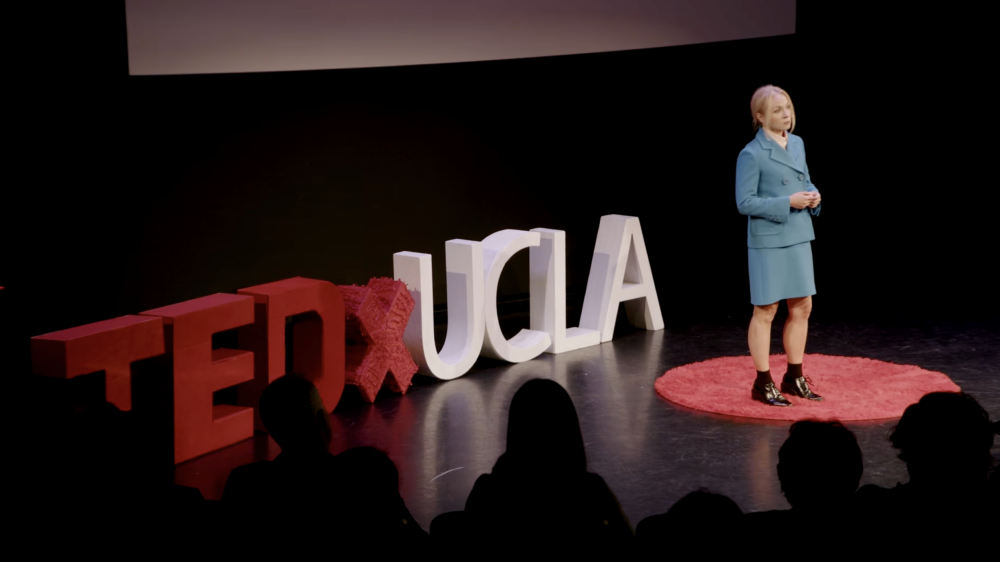 Kelly Turner TedXUCLA talk screenshot with her on a black stage in a blue blazer and skirt.