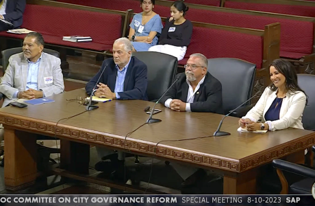 three men and one women at large wooden desk with microphones