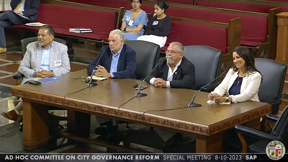 three men and one women at large wooden desk with microphones