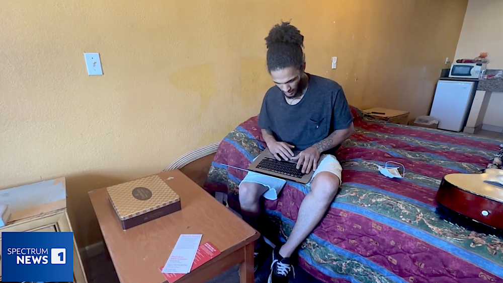 Man on bed in hotel room, typing on laptop