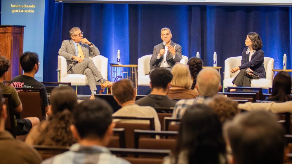 3 panelists speak in front of an audience on stage.|||
