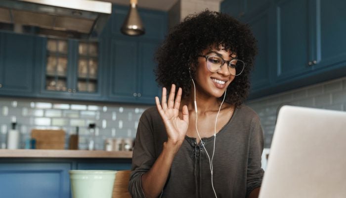 Woman raises hand to introduce herself on video call in front of her laptop sitting in her kitchen.