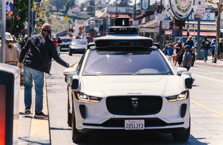 Waymo driverless taxi stops on the street while a passenger reaches out to open the door.