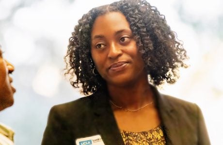 Young Black woman wearing a dark jacket and gold top looks at a person to her right while they chat|two Black women pose in front of a poster about a research project|young woman in blue jacket gestures while talking about research shown on poster to her right