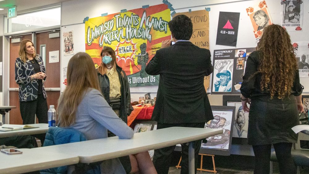 people stand in front of a wall covered with posters, photos and artifacts relating to homelessness