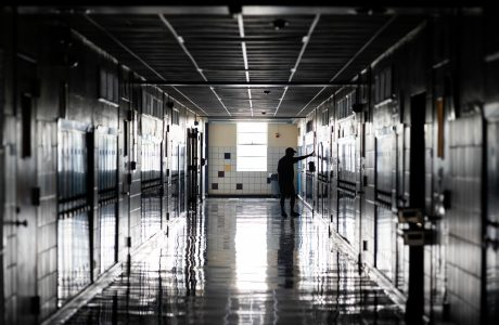 Dimly lit hallway of a high school, with lockers on either side, a man stands with his arms out touching a wall, conveying sadness.