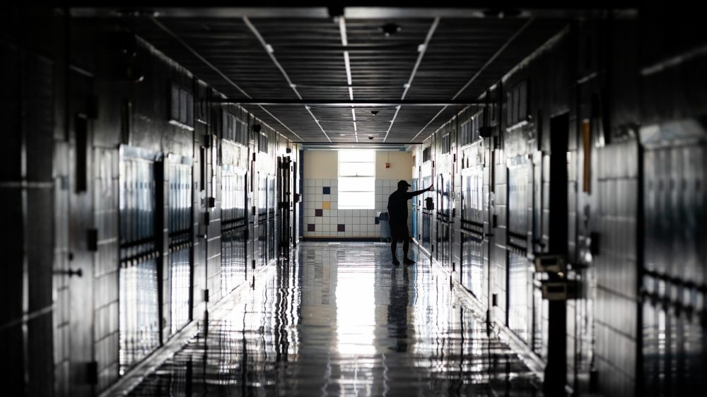 Dimly lit hallway of a high school, with lockers on either side, a man stands with his arms out touching a wall, conveying sadness.
