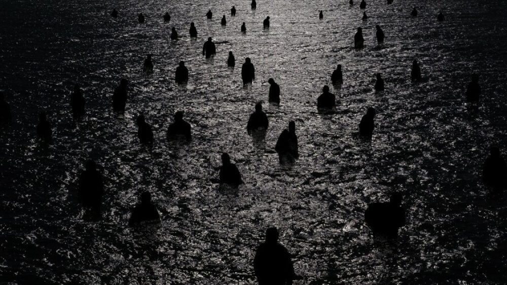 Many dark figures wade in the moonlit water in a black-and-white photo.