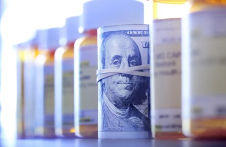 A roll of one hundred dollar bills sits among a row of prescription medication bottles.|Headshot of man in suit, white shirt, tie