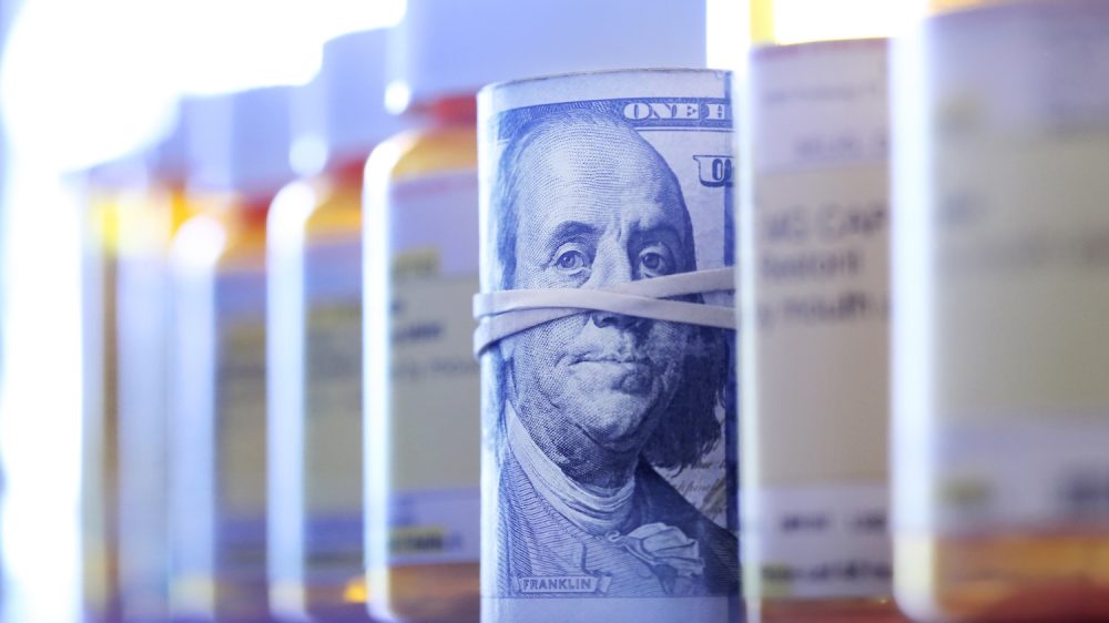 A roll of one hundred dollar bills sits among a row of prescription medication bottles.|Headshot of man in suit, white shirt, tie