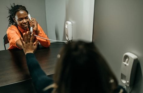 Female prisoner in an orange jumpsuit holds her hand up to the plastic divider while talking to her sister on the other side.