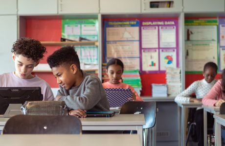 Multiracial group of adolescent kids sit at desks in their classroom and use tablet computers. Selective focus on two boys who are working on an assignment together.