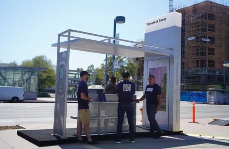 Three people stand underneath a bus shelter on a sunny day.