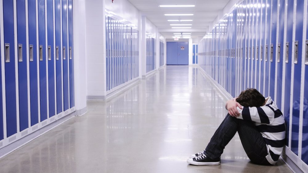 boy sitting with head in hands in school corridor amid blue lockers